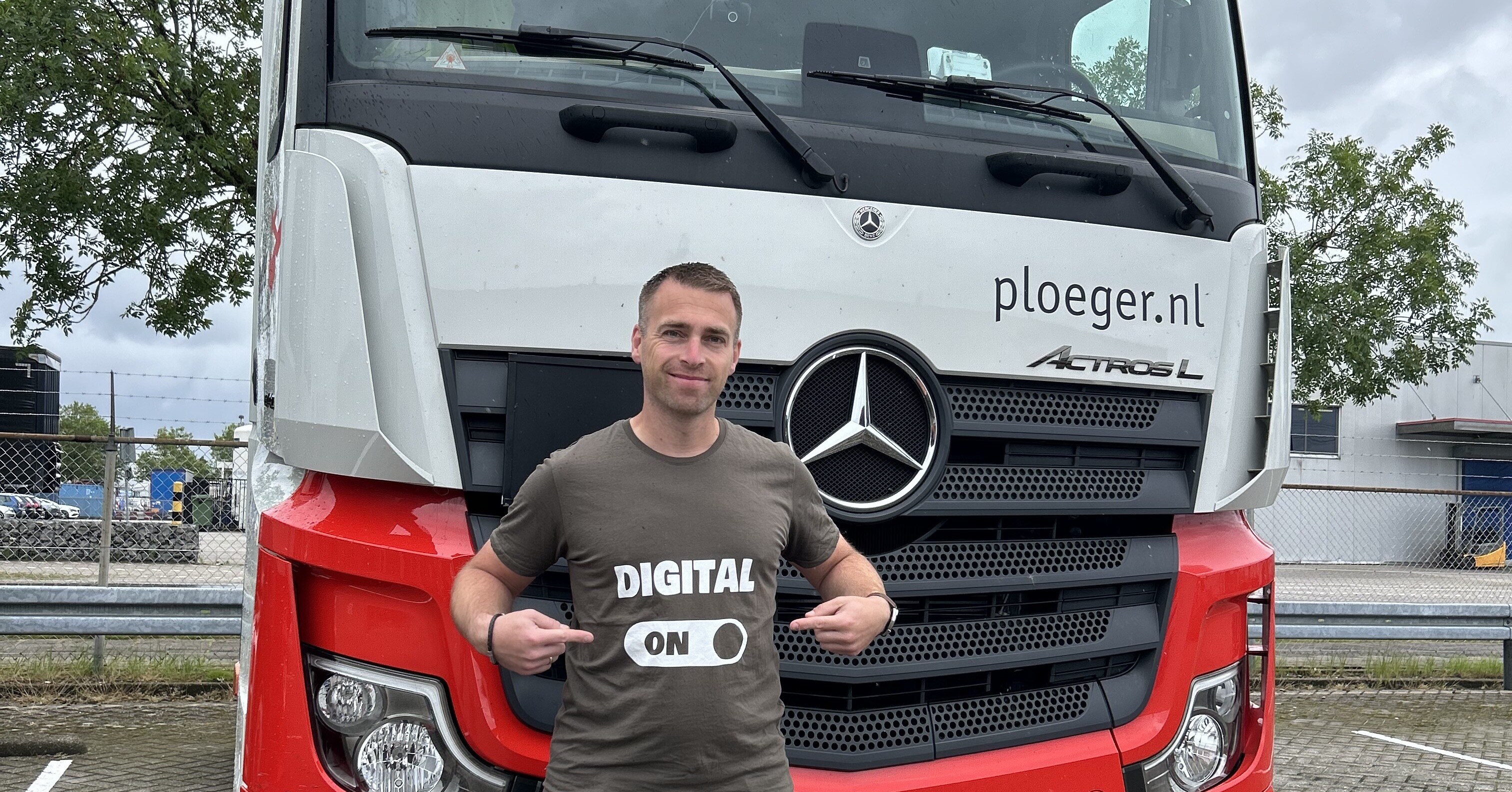 Een man voor een vrachtwagen van het merk Mercedes, te zien aan de voorkant van de wagen. De man wijst naar zijn T-shirt, waarop staat 'Digital On'